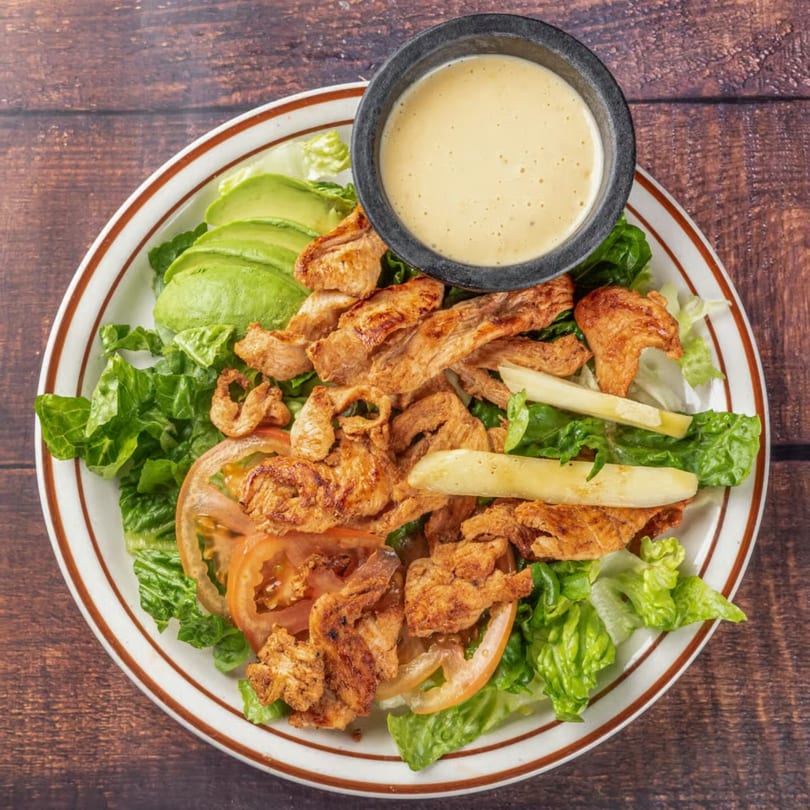 Grilled chicken salad with avocado, tomatoes, and a side of creamy dressing on a wooden table.